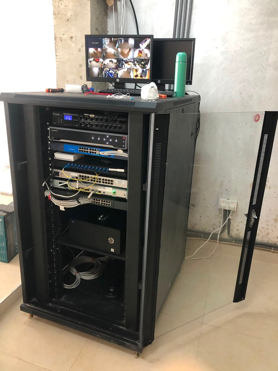 KENVO network and CCTV cabinet with recorder, switches and fibre patching, beside a live multi-camera monitor